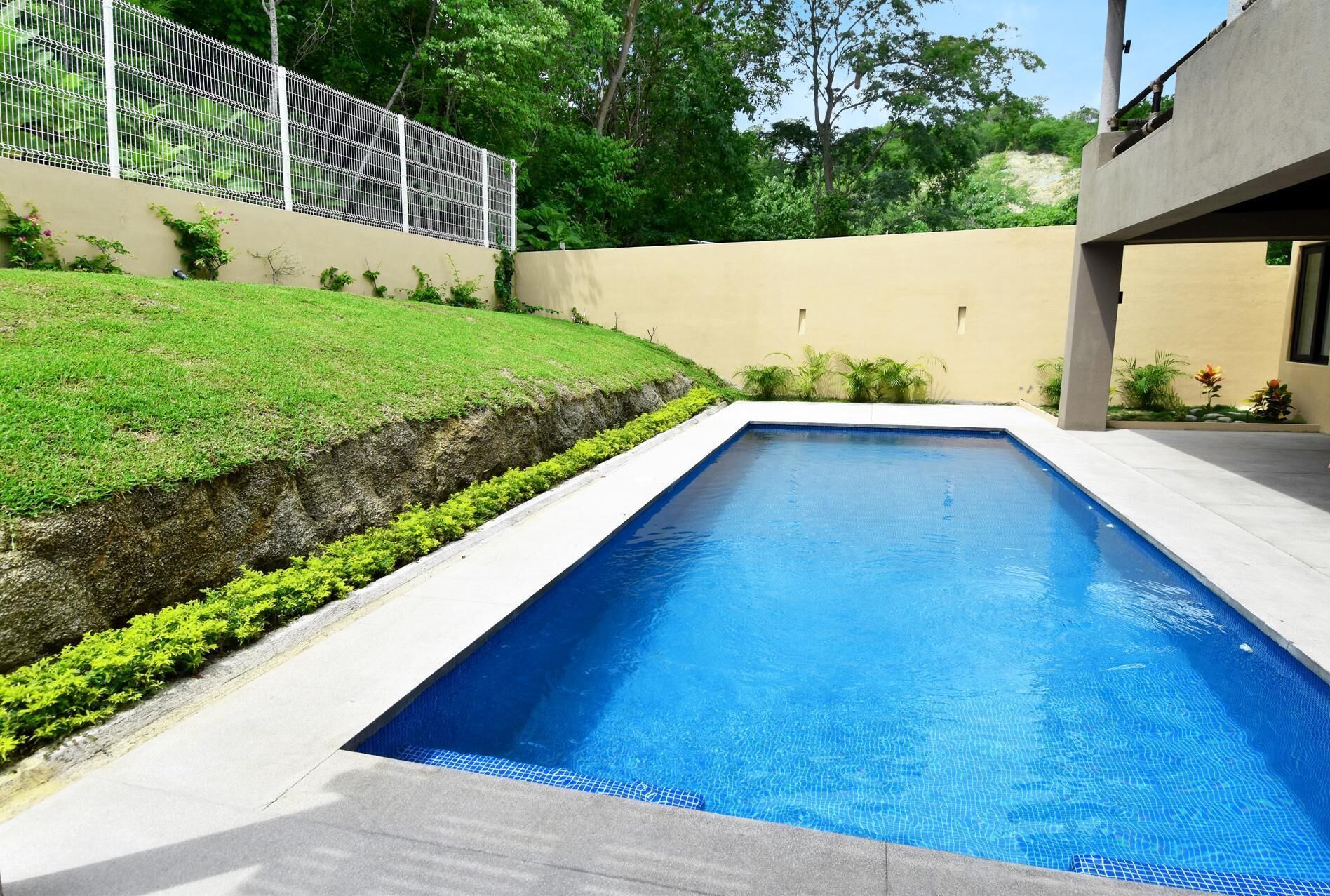 Una piscina rectangular con agua azul cristalina, rodeada por un borde de hormigón y un muro bajo de hormigón.