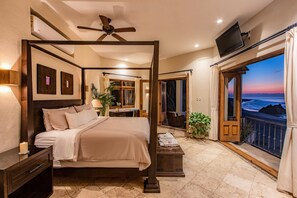 A bedroom with a four-poster bed, a TV mounted on the wall, and a balcony with a view of the sea.