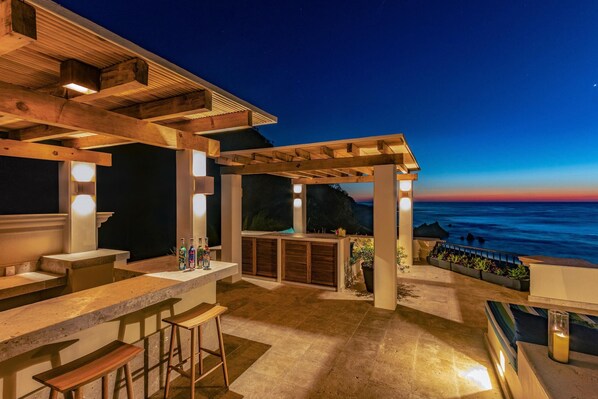 An outdoor bar area with a view of the ocean at dusk, wooden pergola, and bar stools.