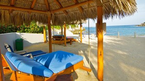 Eine Strandlounge mit einem Reetdach, zwei blaue Liegestühle und Blick aufs Meer.