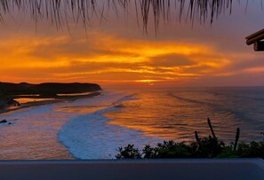 Una vista al atardecer sobre una playa con olas, una costa lejana y algo de vegetación en primer plano.