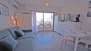 A modern apartment with a grey sofa, a dining table with chairs, a TV, and a balcony with a view.