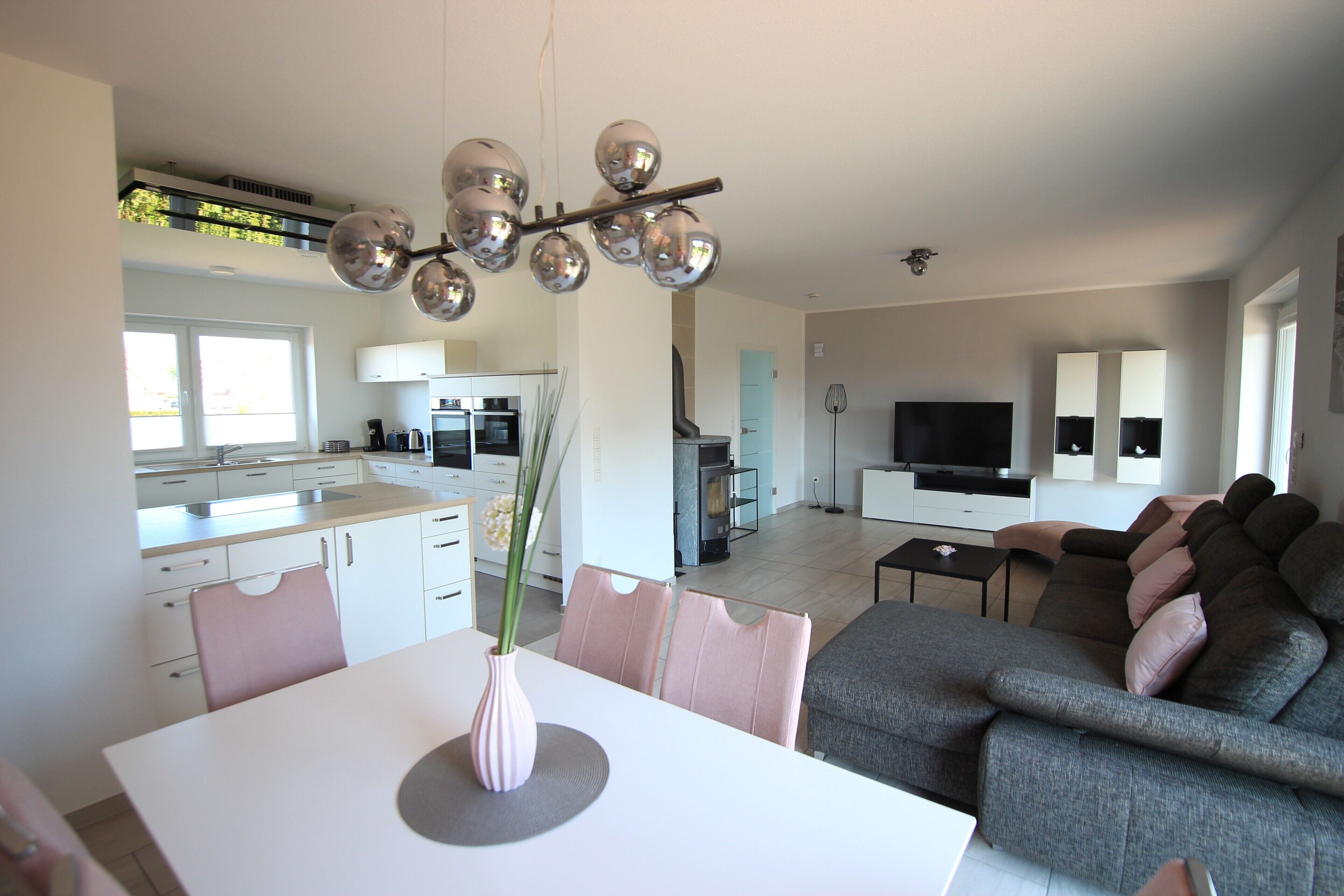A modern kitchen with white cabinets, a dining table with pink chairs, a grey sofa, and a TV in the living area.