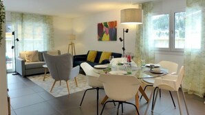 En moderne stue med spiseplads, med en sofa, stole, et spisebord dækket til fire personer, og et farverigt abstrakt billede på væggen.