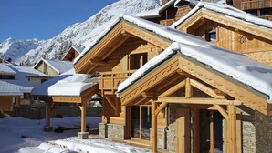 A wooden chalet with a sloped roof covered in snow, set against a mountainous backdrop.