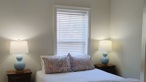 Une chambre à coucher comprenant un lit, deux tables de chevet avec des lampes, un ventilateur de plafond, une fenêtre avec des stores et une porte.