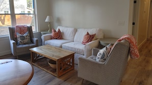 Un salon avec un canapé blanc, deux fauteuils, une table basse en bois et une petite table d’appoint avec une lampe.