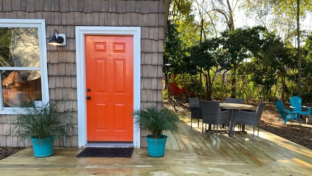 Une maison avec une porte rouge, une terrasse en bois et deux plantes en pot.