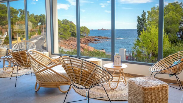 A room with large windows overlooking a coastal landscape, wicker furniture, and a small table with a menu. - Appart Abordable pour 4 | Accès Piscine (Saint-Raphaël)