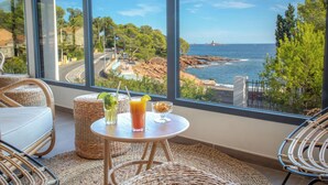 A balcony with wicker furniture, a table with drinks, and a view of the sea and rocky coastline. - Appart Abordable pour 4 | Accès Piscine (Saint-Raphaël)