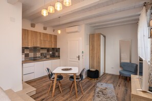 A modern kitchen with wooden cabinets, a tiled backsplash, and a dining area with a table and chairs. There is a black ottoman and a blue chair in the living area.
