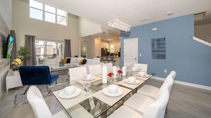 A modern dining area with a glass-top table set for six, white chairs, and a blue accent chair. A chandelier hangs from the ceiling, and a kitchen is visible in the background. - SG2764 Storey lake  5 bed 5 bath Private Pool & Free Waterpark (Kissimmee)