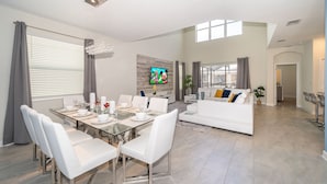 A modern living room with a dining area, white furniture, a flat-screen TV, and a large window. - SG2764 Storey lake  5 bed 5 bath Private Pool & Free Waterpark (Kissimmee)