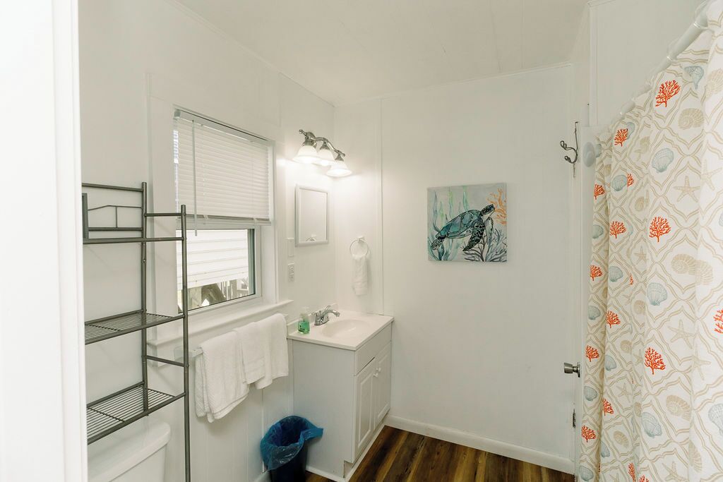 A compact bathroom with a toilet, sink, and shower area. There are shelves on the left, a towel rack, and a trash bin.