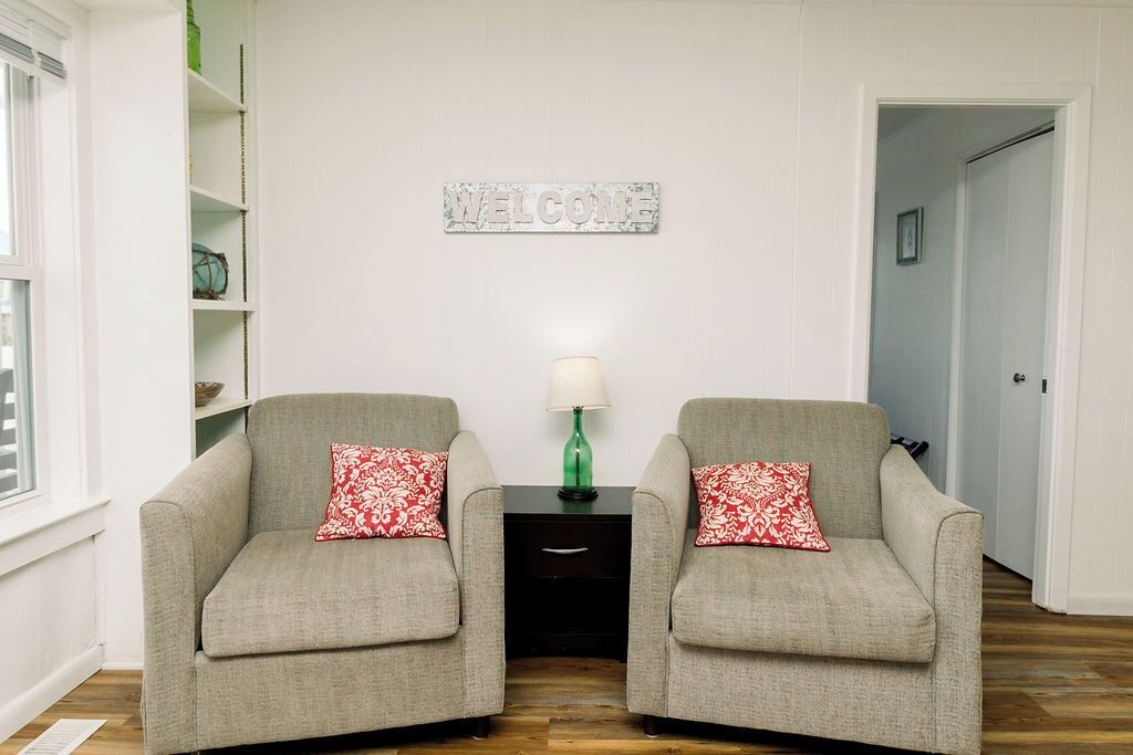 A room with two grey armchairs, a small table with a green lamp, and a 'WELCOME' sign on the wall.