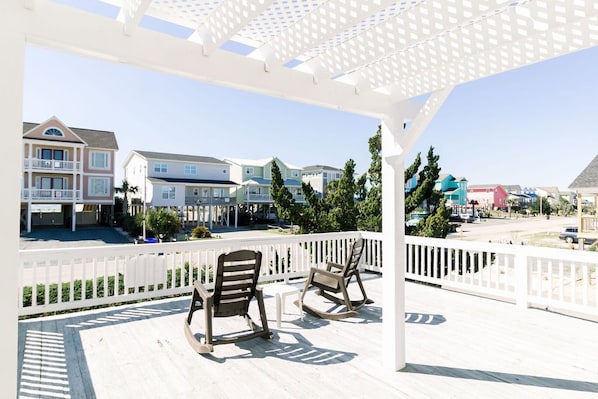 A covered outdoor deck with two rocking chairs, a white fence, and a view of multi-story houses.