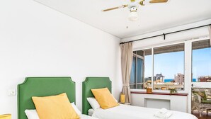 Ett hotellrum med två sängar, ett stort fönster med utsikt över byggnader och havet, en takfläkt och ett litet bord med en lampa.