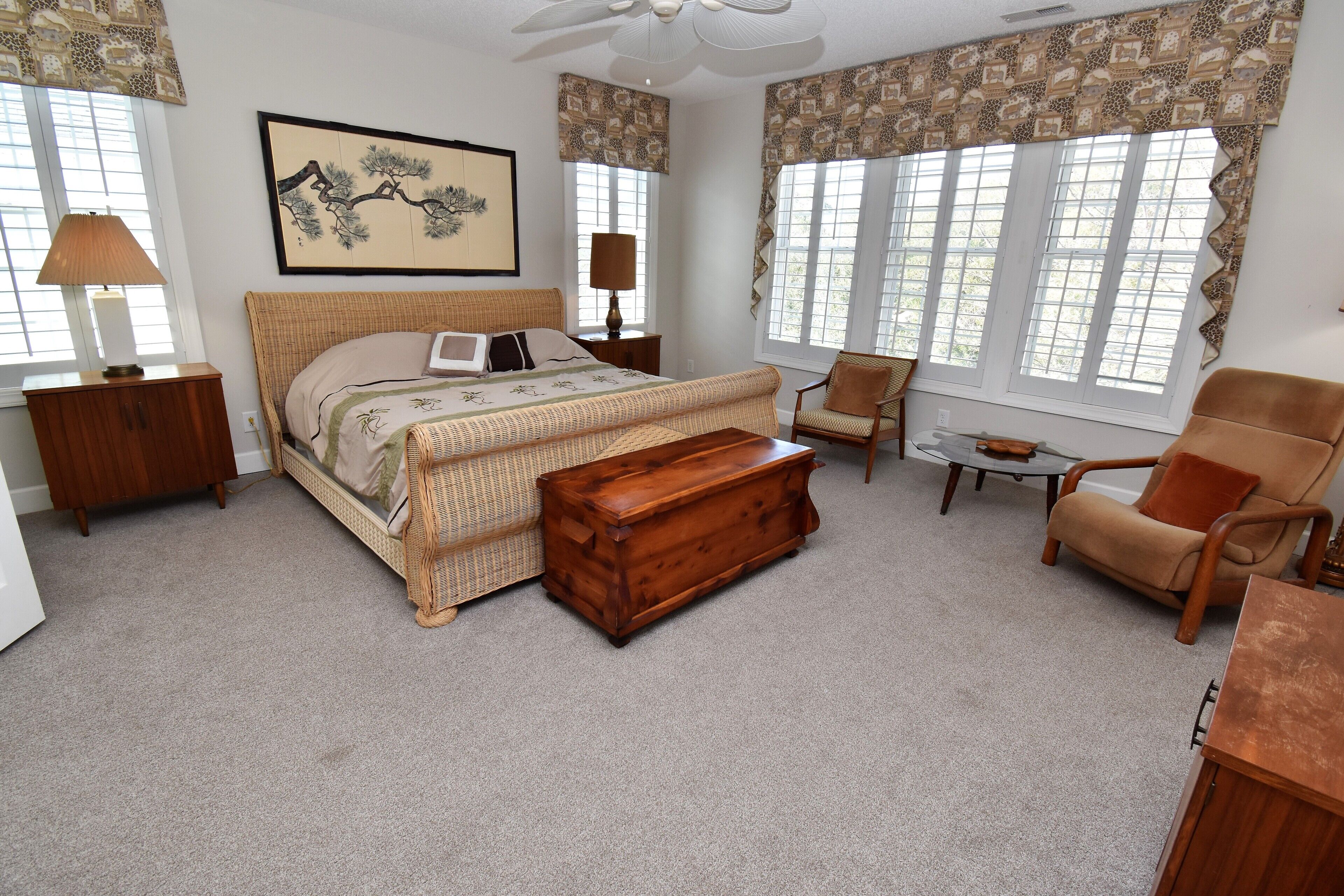 A bedroom with a large bed, a wooden chest, a chair, a small table, and a ceiling fan.