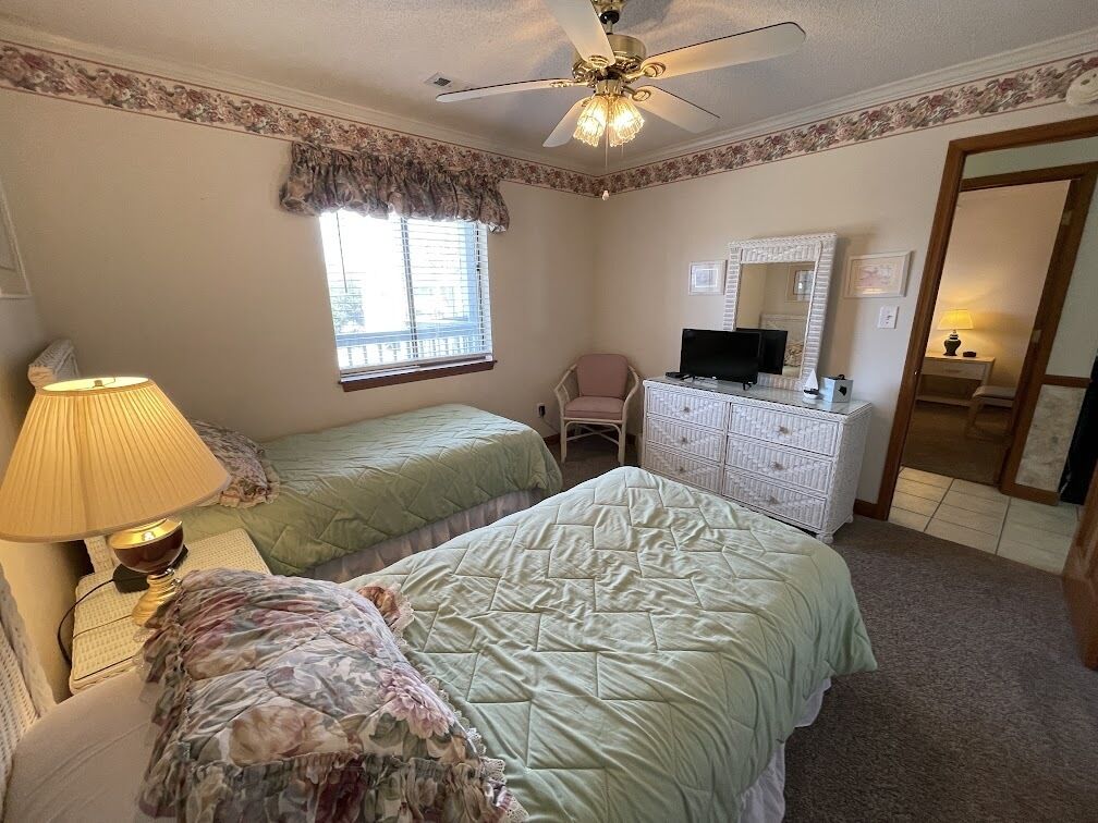 A bedroom with two beds, a ceiling fan, a dresser, a television, and a window with blinds.