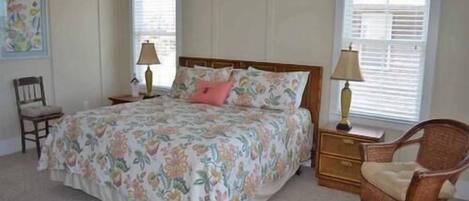 Un dormitorio con colcha estampada, cabecero de madera, dos lámparas de noche, una silla de mimbre y un cuadro enmarcado en la pared.