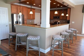 A kitchen with wooden cabinets, a stainless steel refrigerator, a granite countertop, and bar stools with patterned cushions.