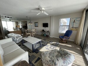 A spacious living room with a ceiling fan, a dining area with a table and chairs, a blue armchair, and a large window with blinds.