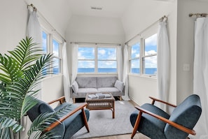 A room with a sofa, two armchairs, a coffee table, and a large window with a view of the sea.