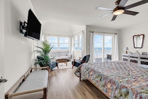 A bedroom with a bed, a TV, a ceiling fan, a balcony with a view, and a potted plant.