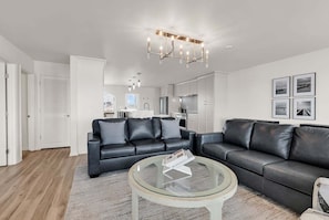 A modern living room with two black leather sofas, a glass coffee table, and a kitchen in the background.