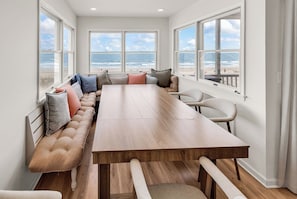 Una habitación amplia con una larga mesa de madera, asientos acolchados y ventanas grandes con vistas al océano.