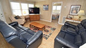 A living room with leather sofas, a flat-screen TV, wooden panel walls, and a large window. - Electa's Place (Up) - Second Row in Garden City Beach (Garden City Beach)