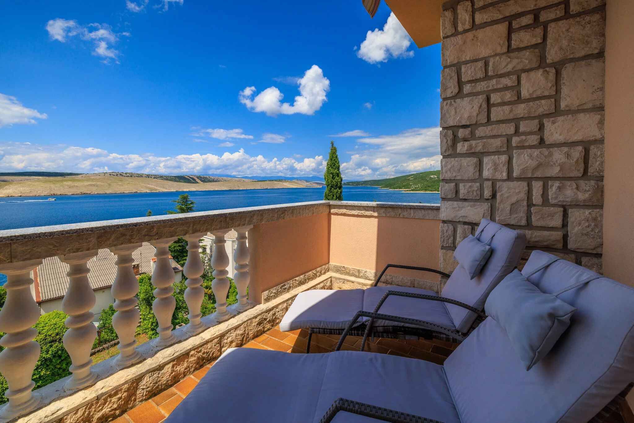 A balcony with a view of a lake and distant hills, furnished with white lounge chairs and a stone wall.