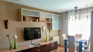 Een moderne woonkamer met een flatscreen televisie, houten kasten, een eethoek met een tafel en stoelen, en een groot raam met zonwering.