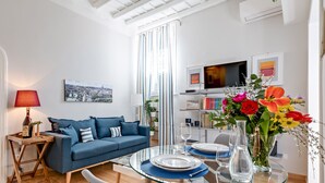 Un salon moderne avec une table à manger en verre pour deux personnes, un canapé bleu, un téléviseur et une grande fenêtre avec des rideaux.