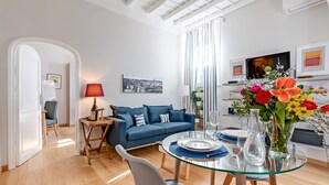 Un salon moderne avec un canapé bleu, une table à manger en verre et un tableau représentant un paysage urbain accroché au mur.