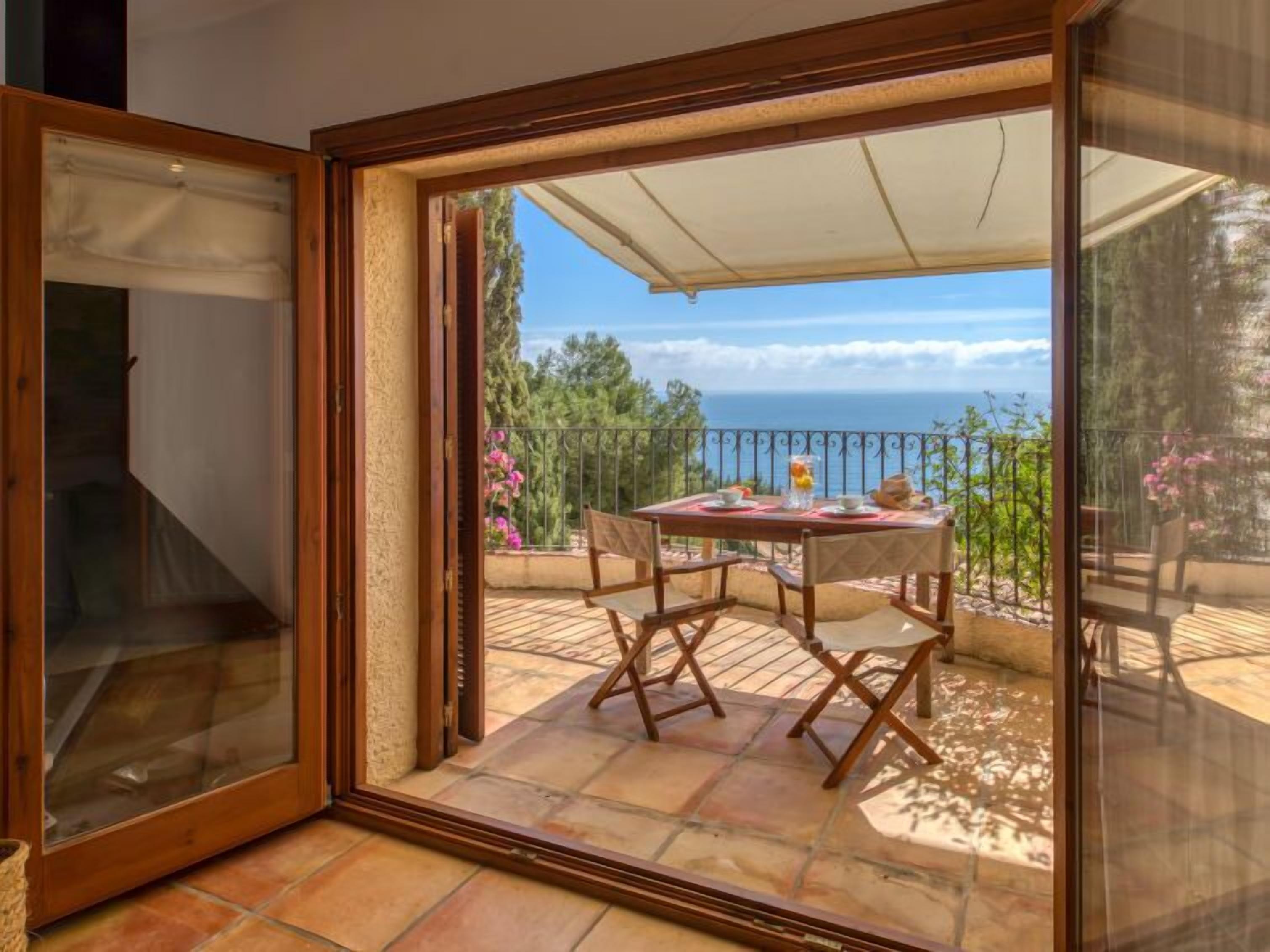 A room with a sliding glass door leading to an outdoor patio with a table set for two, overlooking a scenic view of the sea.