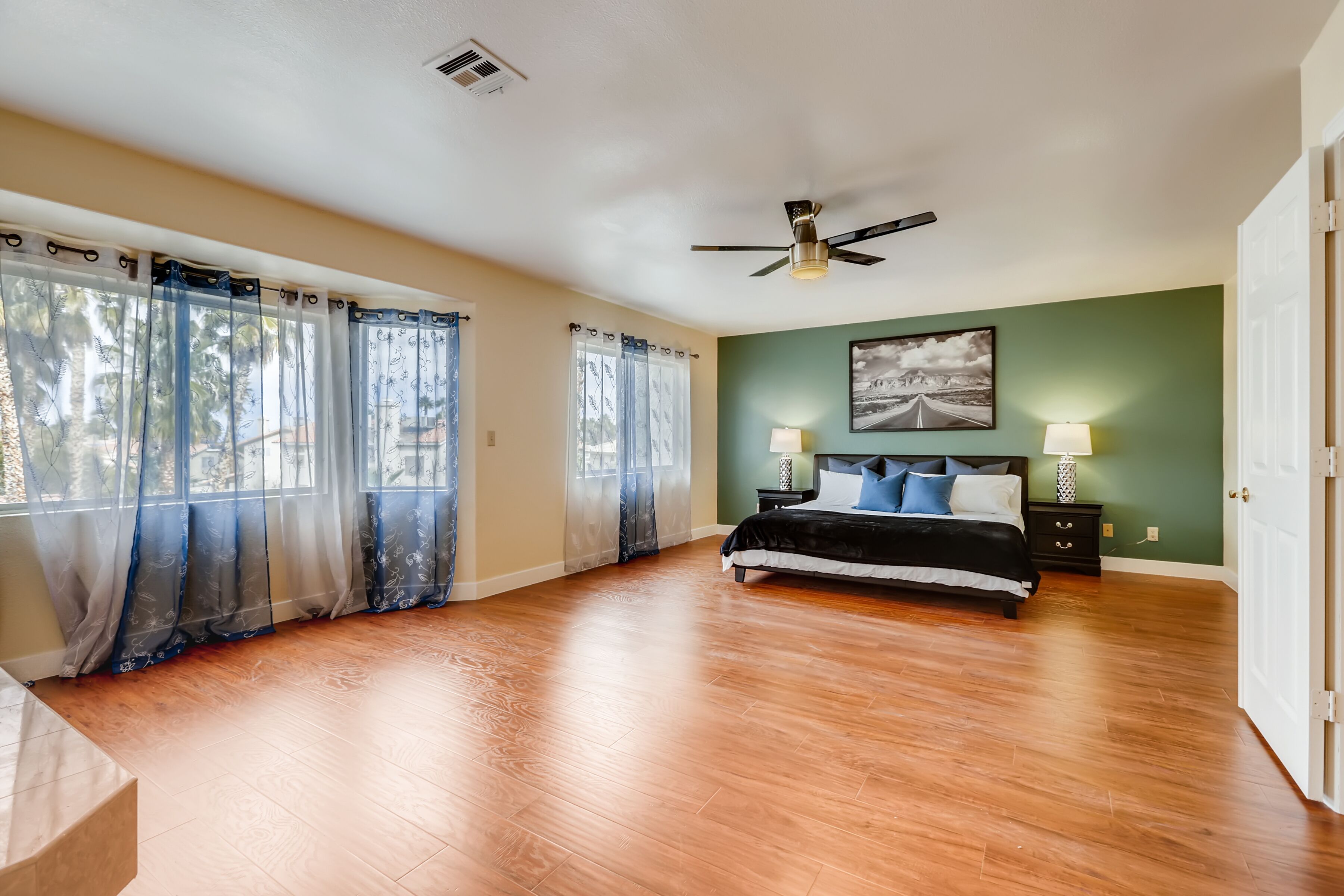 A spacious bedroom with a large bed, wooden flooring, a ceiling fan, and two bedside lamps.