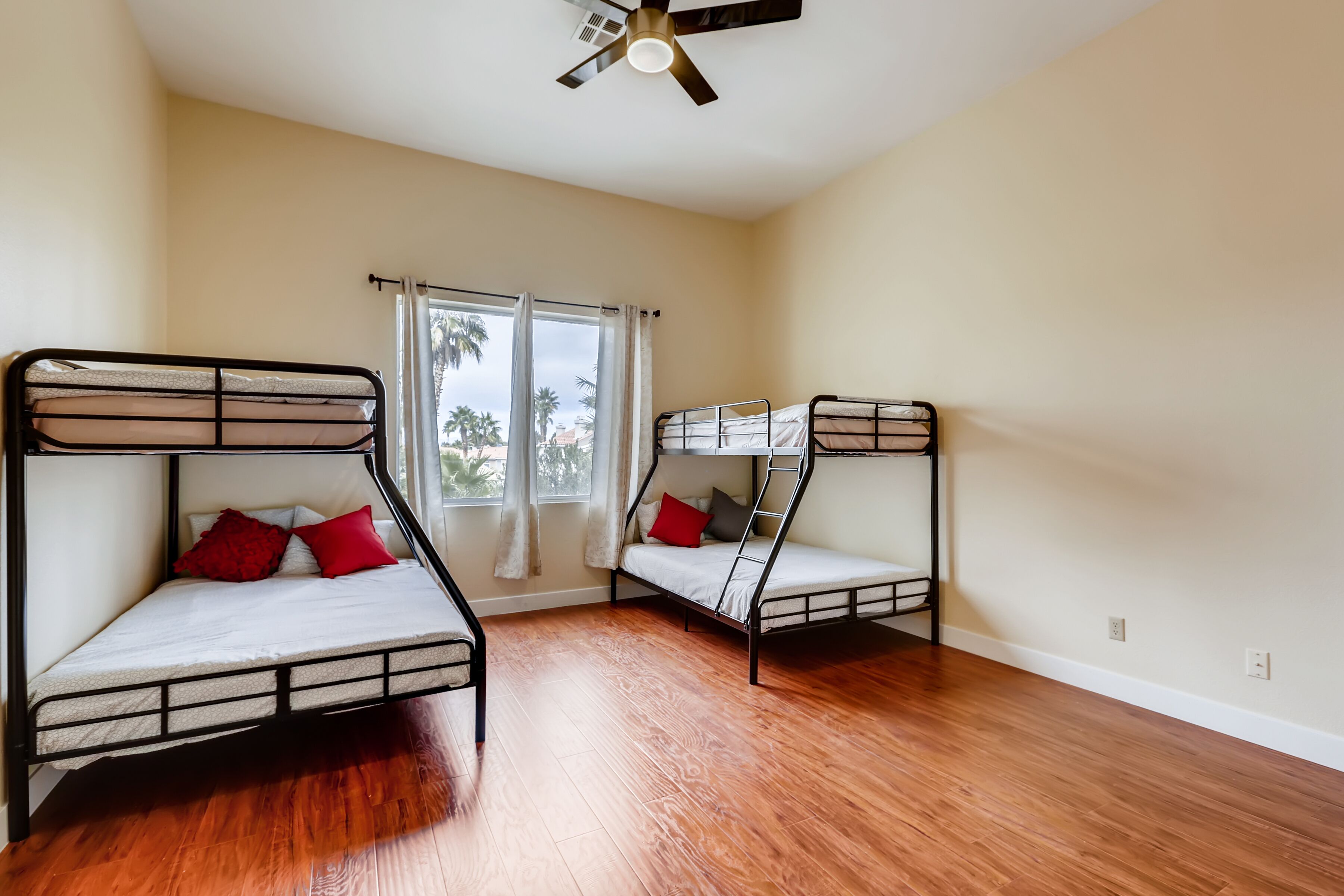 A room with two bunk beds, wooden flooring, a ceiling fan, and a window with curtains.