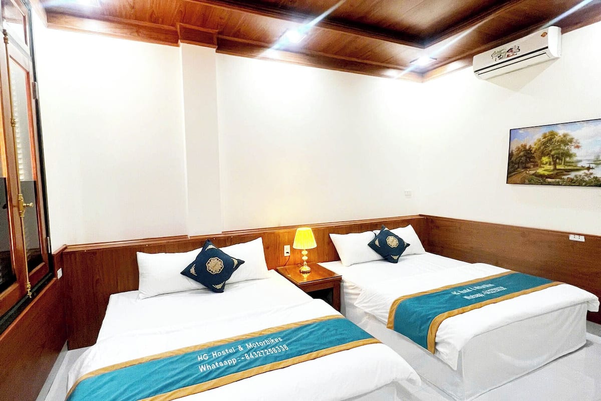 A hotel room with two beds, wooden headboards, a nightstand with a lamp, a painting on the wall, and an air conditioning unit.