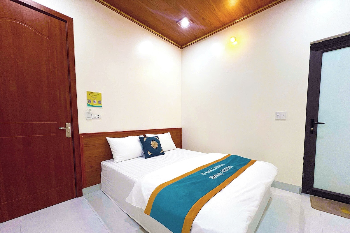A hotel room with a wooden door, a bed with white bedding and a blue and gold bedspread, and a light switch on the wall.