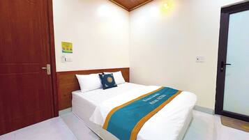 A hotel room with a wooden door, a bed with white bedding and a blue and gold bedspread, and a light switch on the wall.