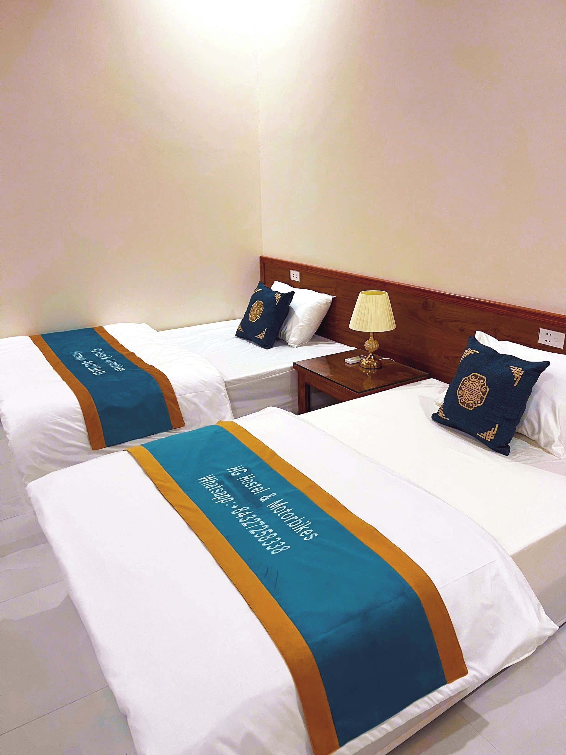 Two single beds with white linens and teal and gold bedspreads, a wooden headboard, a bedside table with a lamp, and a wall socket.