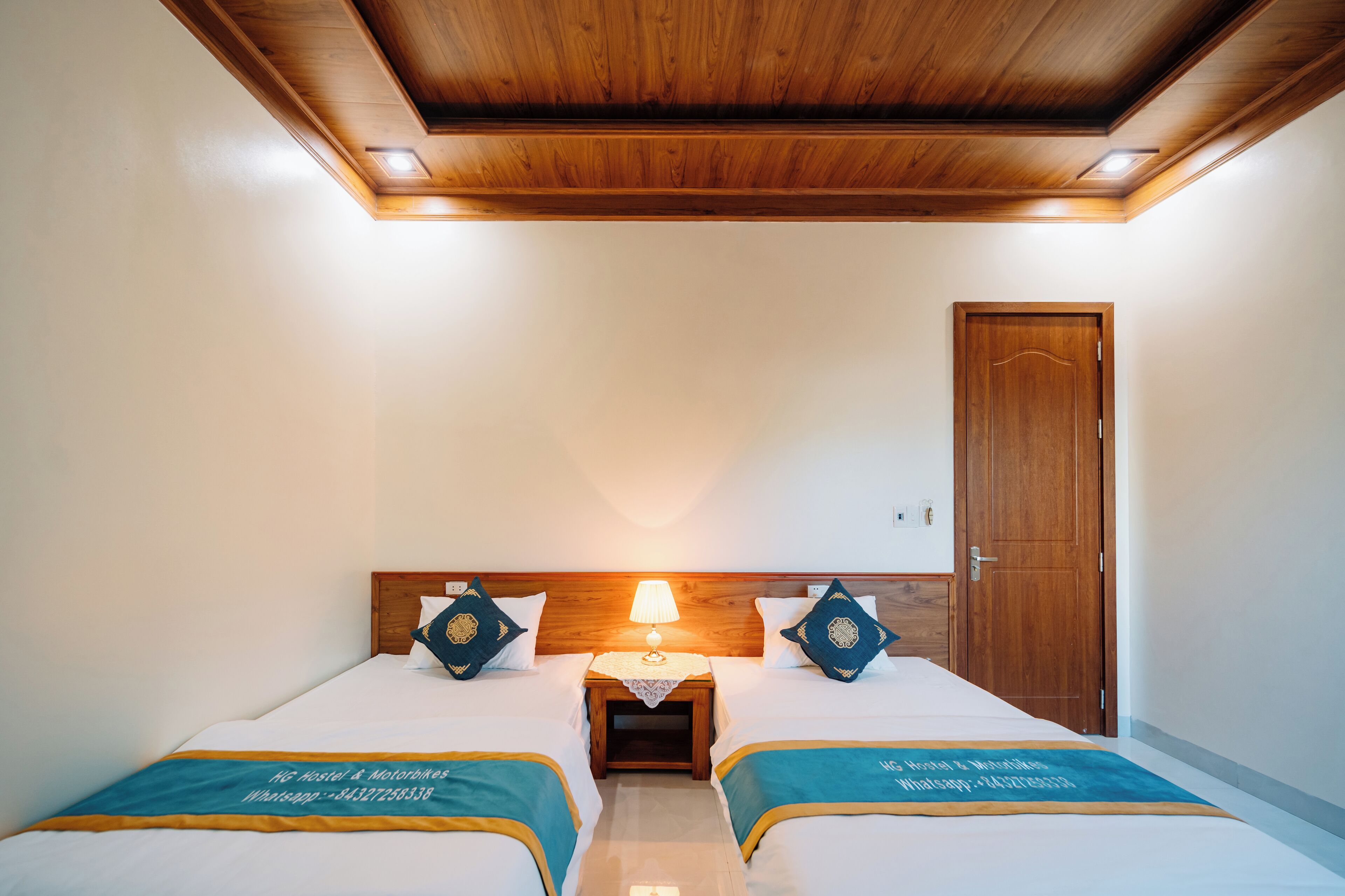 A hotel room with two beds, a wooden ceiling, a nightstand with a lamp, and a door.