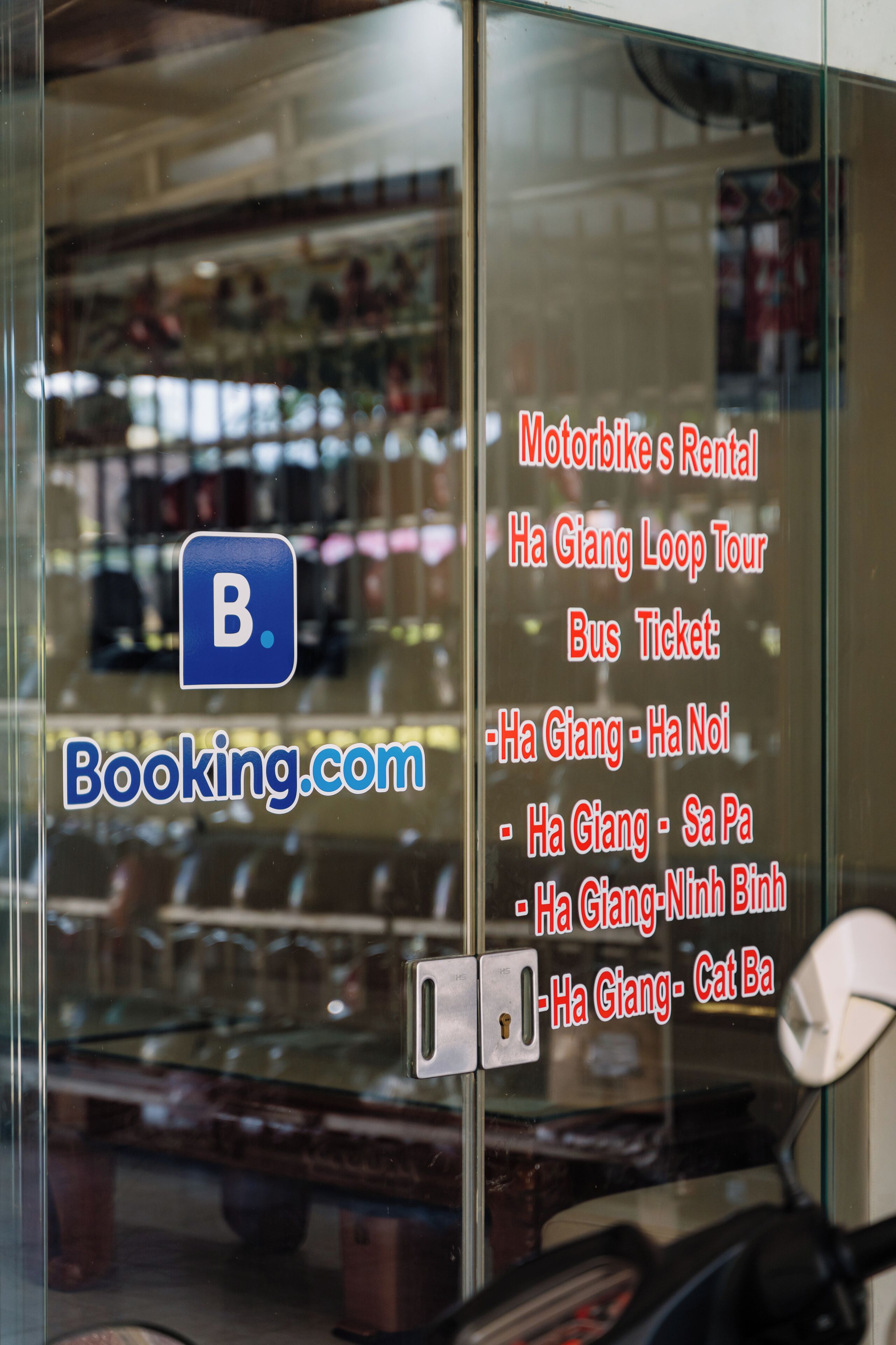 A glass window with a booking.com logo and text advertising motorbike rentals and bus tickets for Ha Giang Loop Tour.
