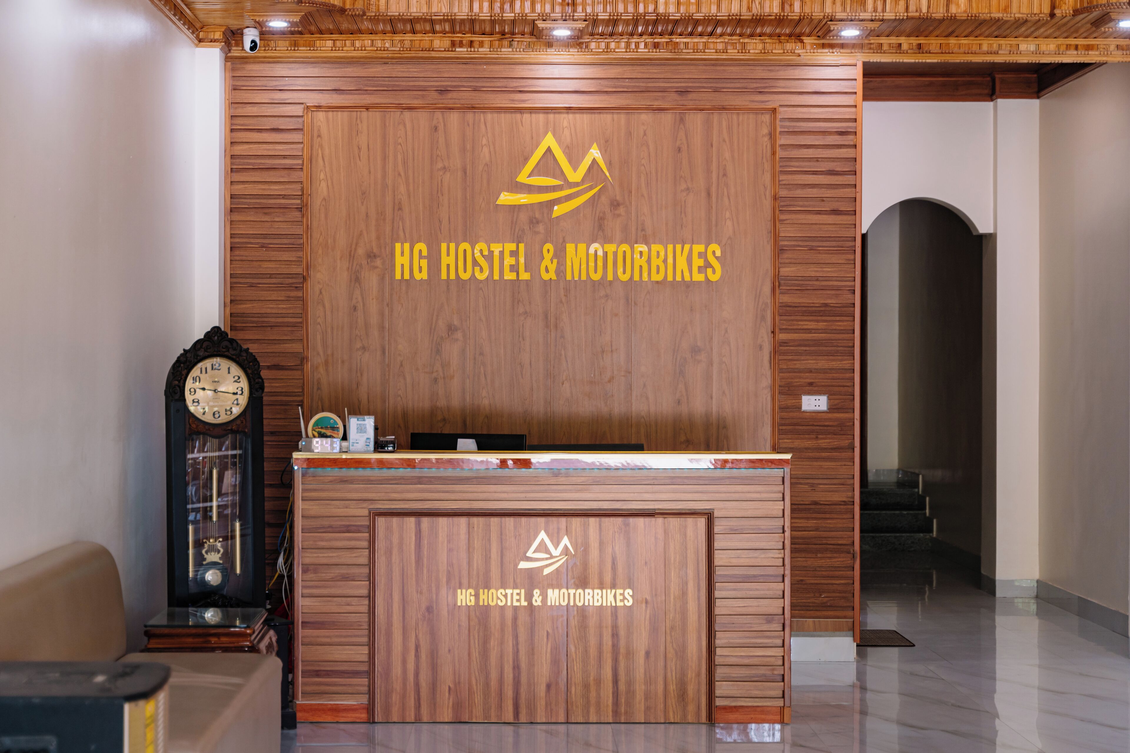 A reception desk with a wooden finish and the logo "HG HOSTEL & MOTORBIKES" on the wall behind it.
