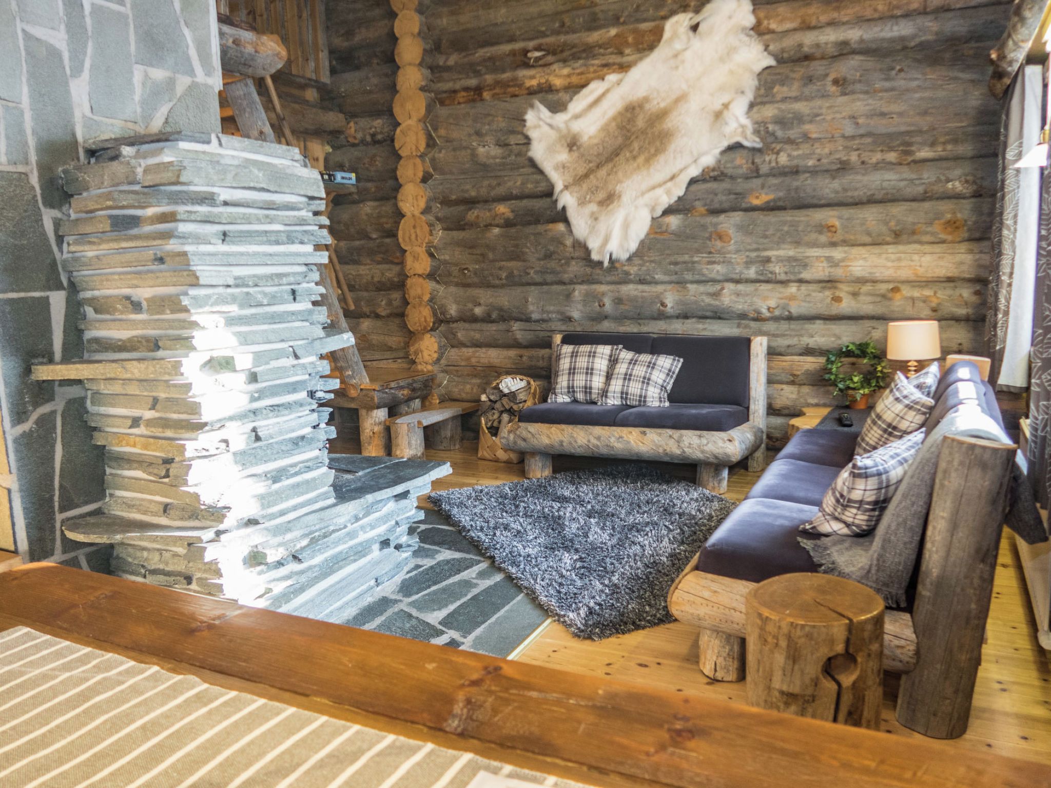 Ein Blockhaus-Innenraum mit Kamin aus Stein, einer Couch mit Karokissen und einem Fellteppich.
