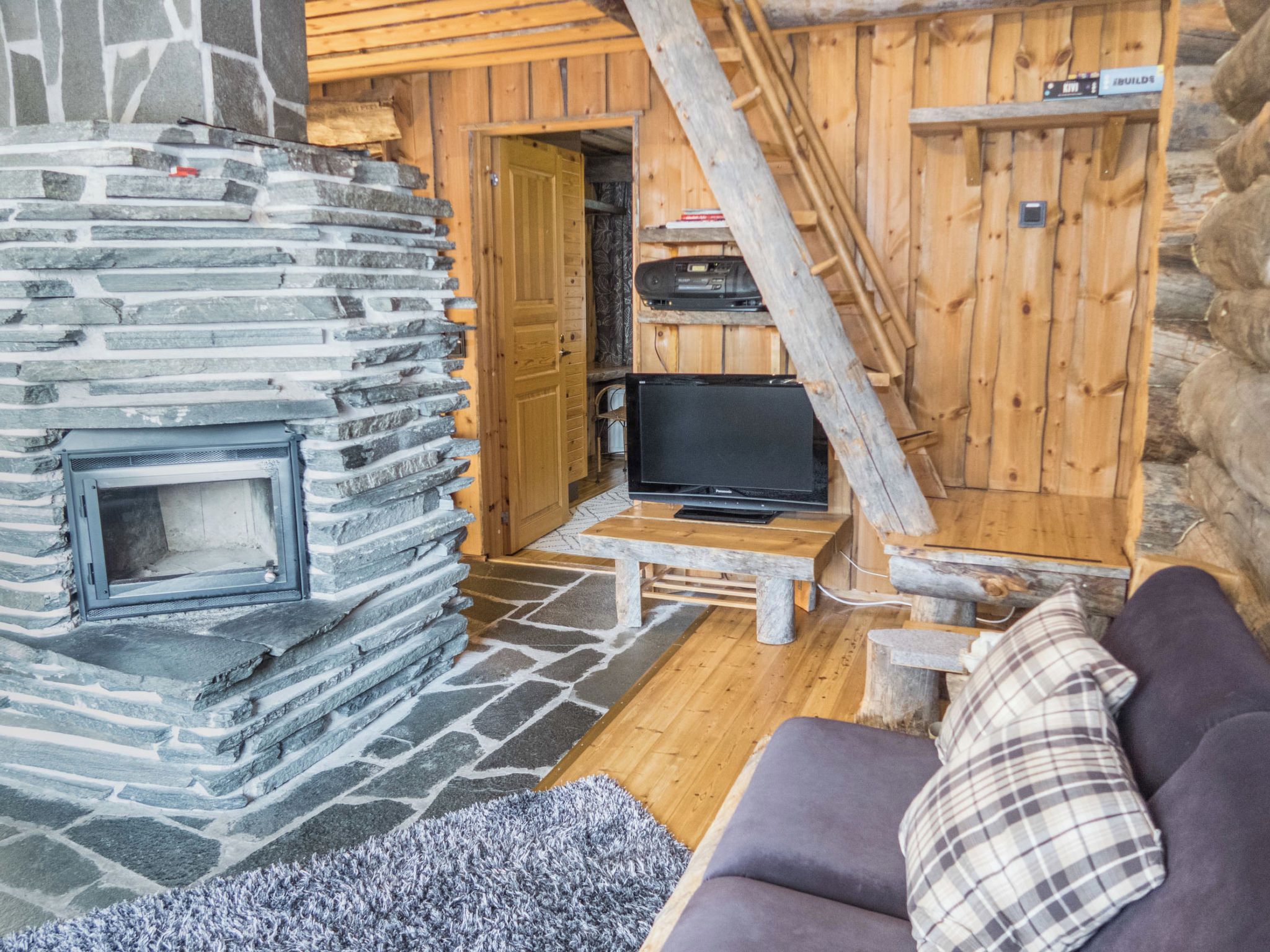A cozy cabin interior with a stone fireplace, a television on a wooden table, and a couch with a plaid pillow.