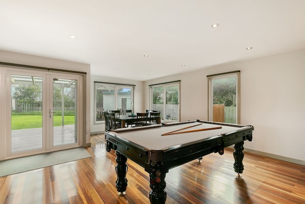 Une pièce spacieuse comprenant une table de billard, un coin repas avec des chaises et de grandes portes-fenêtres coulissantes donnant sur un jardin.