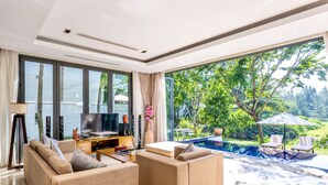 A modern living room with a large TV, sofas, a coffee table, and a view of a swimming pool and greenery.