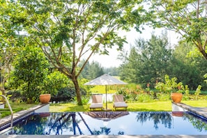 An outdoor swimming pool with two lounge chairs and an umbrella, surrounded by lush greenery and trees.